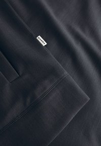 Sötétszürke szövet texturált felülettel, zsebbel és egy fehér "GABBA" feliratú címkével. Tiszta vonalak és minimalista dizájn.