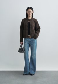 Brown knit cardigan with button closure, two front pockets, paired with flared blue jeans. Accessorized with a small black handbag.