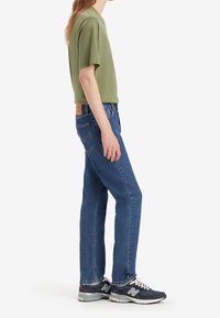 Personne debout portant une chemise à manches courtes vert olive, un jean bleu et des baskets noires New Balance sur fond blanc uni.