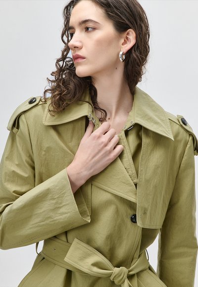 Γυναίκα με καστανά σγουρά μαλλιά, φοράει πράσινο παλτό τύπου τρεντσκότ με επιχρωμιωμένους ώμους, ζώνη στη μέση και μεγάλα ασημένια σκουλαρίκια κρίκους, που κοιτάζει αριστερά.