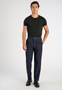 Camiseta preta ajustada combinada com calças escuras de cintura alta e texturizadas, apresentando pregas frontais e um fecho de botão único. Sapatos pretos.