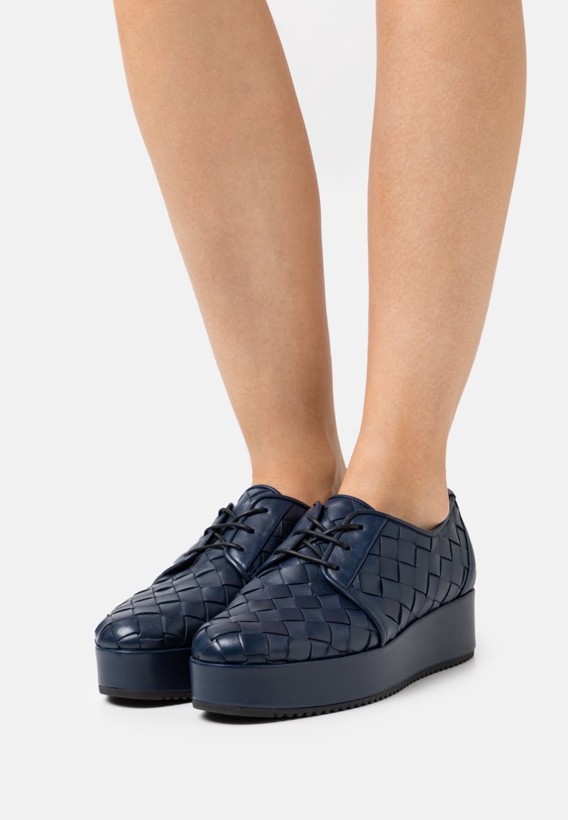 Jambes portant des chaussures à plateforme à lacets en cuir tissé bleu marine avec semelles épaisses sur un fond clair uni.