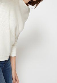 Maglione bianco con texture, collo rotondo e polsini a coste, abbinato a jeans blu scuro. Il maglione presenta un motivo geometrico sottile.