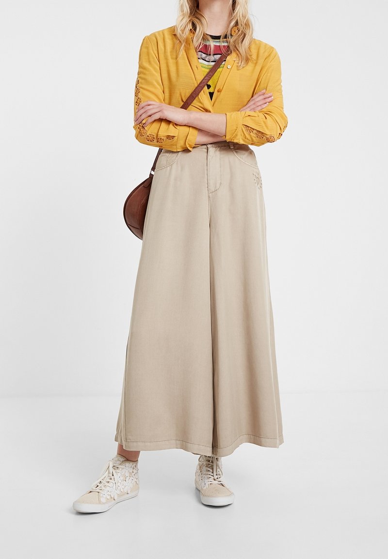 Femme debout, les bras croisés, portant une veste jaune brodée, un pantalon beige à jambes larges, des baskets crème à motifs floraux et un sac bandoulière marron.