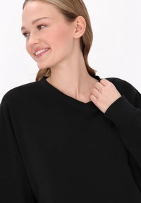 Sweatshirt negru cu decolteu în V, cu mâneci lungi și manșete ribbate; materialul pare moale cu o textură netedă. Mâna modelului atinge gulerul.