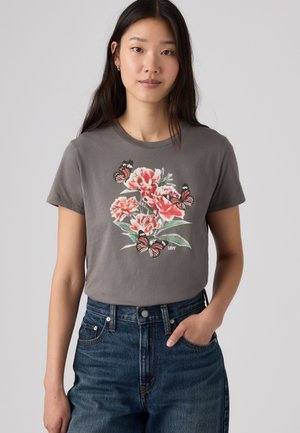 Jeune femme portant un t-shirt gris avec un imprimé fleur rose et papillon, associé à un jean bleu taille haute sur un fond uni.