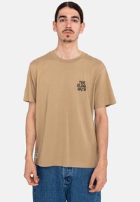 Beige cotton t-shirt with short sleeves, round neckline, and black printed text on the left chest. Worn with blue denim shorts.