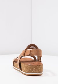Brune semsket sandaler med en flat, korkstrukturert plattform, justerbar ankelrem og beige polstret såle. Bakvinkel med fokus på reimdesignet.