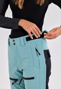 Light blue soft-shell pants with a black accent, featuring a zip fly, snap buttons, and side pockets with zippers. Velcro strap at the waist.