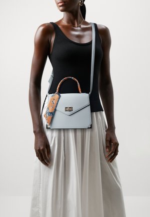 Frau wearing ein schwarzes ärmelloses Oberteil und einen weißen Rock, die eine hellblaue Handtasche mit einem gemusterten orangefarbenen Schal am Griff hält.