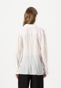 Blusa bianca con pieghe, caratterizzata da un colletto a punta, maniche lunghe e polsini con bottoni. Il tessuto è semitrasparente con righe verticali.
