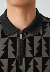 Pulover negru și bej cu fermoar, având un model geometric de forme în zigzag și un guler texturat.