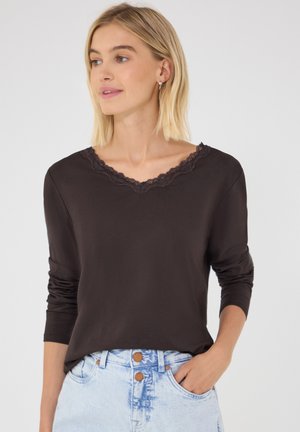Blondynka w czarnej bluzce z długim rękawem ozdobionej koronką i jasnoniebieskich dżinsach z wysokim stanem, z jedną ręką w kieszeni, patrząca w lewo.