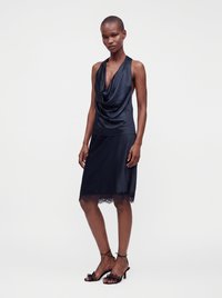 Femme portant une robe noire en satin sans manches avec un décolleté drapé et un ourlet en dentelle, associée à des sandales noires à talons hauts et à brides.