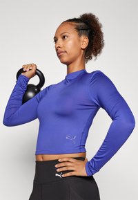 Blue long-sleeve athletic shirt made of smooth fabric, featuring a fitted design and a small logo on the left side. Black leggings complement it.