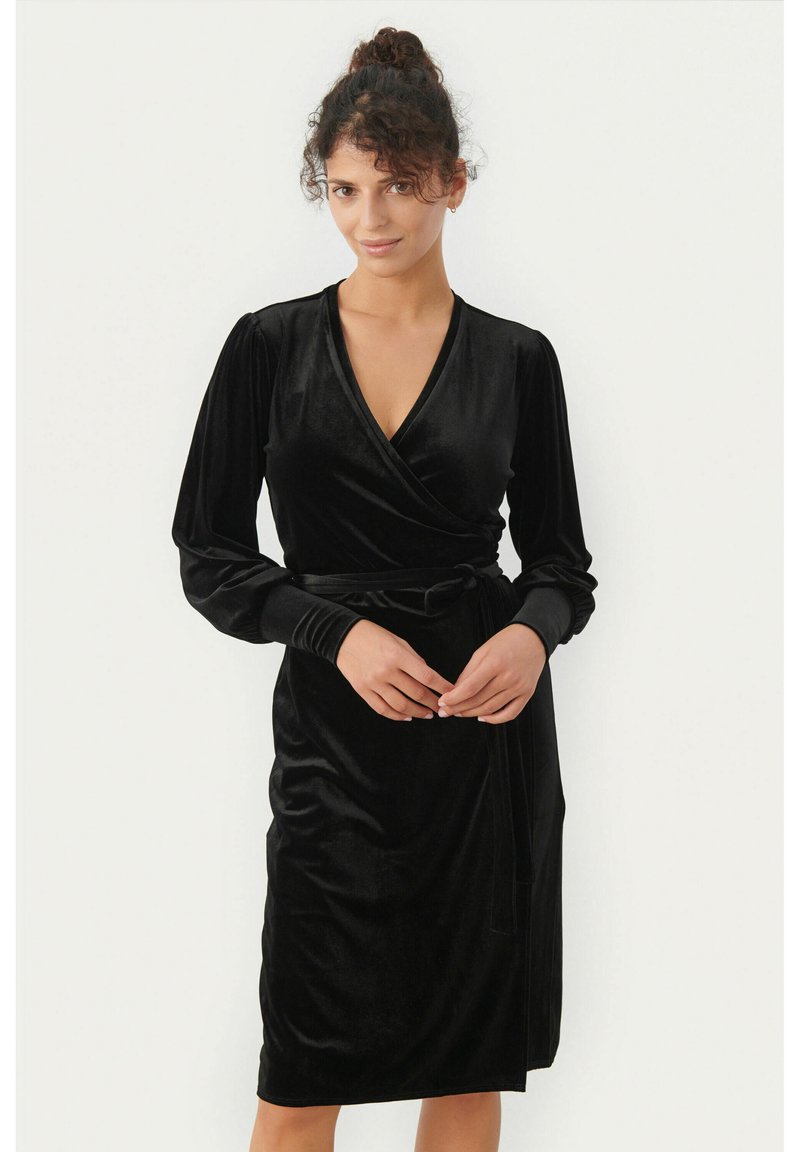 Vestido de terciopelo negro con mangas largas abullonadas, escote en V y cintura anudada. Textura suave y diseño hasta la rodilla.