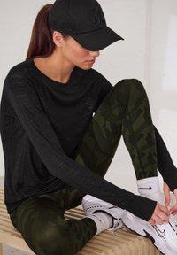 Schwarzes langärmliges Oberteil mit feiner Textur, grüne Camouflage-Leggings und weiße Sneaker mit schwarzen Akzenten, kombiniert mit einer schwarzen Mütze.
