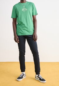 Grön bomullströja med korta ärmar och vitt logotyp, i kombination med svarta smala byxor och svarta sneakers med rutigt mönster.