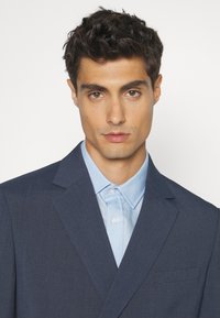 Dunkelblazer mit Revers und einer einzelnen Brusttasche, über einem hellblauen Hemd mit Knopfleiste geschichtet. Glatte Stofftextur.
