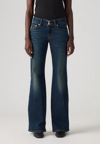 Jean en denim bleu foncé taille haute avec jambes évasées porté avec des chaussures pointues noires et un haut noir sur un fond uni.