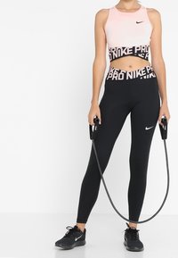 Leggings noirs et un soutien-gorge de sport rose, avec une bande logo, présentant une texture lisse et un design ajusté. La personne tient une corde à sauter.