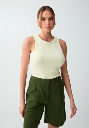 Jeune femme avec un haut beige clair sans manches et un short taille haute vert foncé, debout devant un fond clair uni.