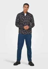 Noor mees seisab käed ristis, seljas must särk, millel on kommitõllide muster, sinised teksad ja mustad mokasiinid, heleda tausta ees.