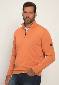 Pullover leggero arancione, realizzato in misto cotone, con mezza zip, polsini e orlo a coste, e una toppa nera sulla manica sinistra.