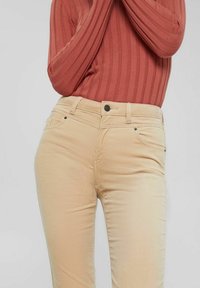 Personne en pantalon beige et un haut à manches longues côtelé couleur rouille, bras croisés devant la poitrine sur fond uni.