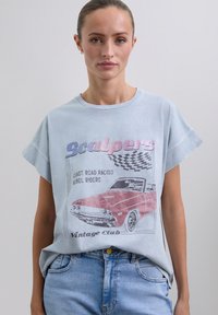 Frau mit einem hellblauen Vintage-T-Shirt, auf dem ein rotes Cabriolet und eine karierten Flagge abgebildet sind, kombiniert mit hellblauen Jeans.