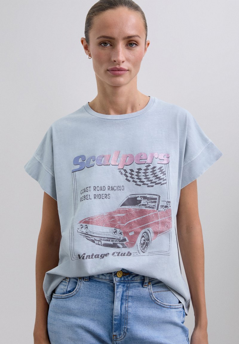 Woman wearing a light blue vintage-style T-shirt with a red convertible car and checkered flag graphic, paired with light blue jeans.