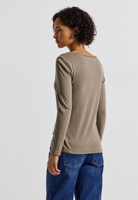 Langärmliges, rundhalsiges Shirt aus beigem Baumwollstoff, körperbetonter Schnitt mit glatter Textur, kombiniert mit blauer Denim, locker sitzende Hosen, sichtbare Nähte.