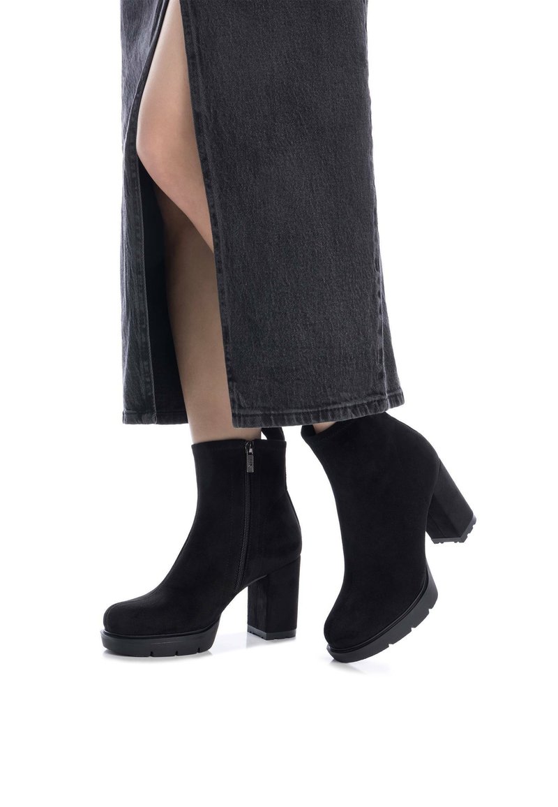 Botas de ante negras hasta el tobillo con tacón grueso, detalle de cremallera y suela texturizada, combinadas con una falda de mezclilla oscura con una abertura lateral.