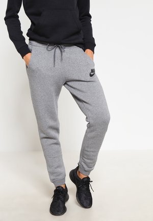 Person wearing grey Nike sweatpants with drawstring, black hoodie, and black athletic shoes, standing with hands in pockets.