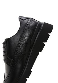 Chaussures en cuir noir avec des détails en brogue, une texture lisse, une semelle épaisse et robuste, et des coutures contrastées le long des bords.