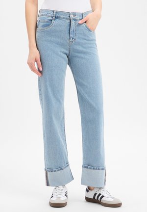 Persoon draagt lichtblauwe opgerolde spijkerbroek met hand in zak en witte sneakers met zwarte strepen, staande op een witte achtergrond.