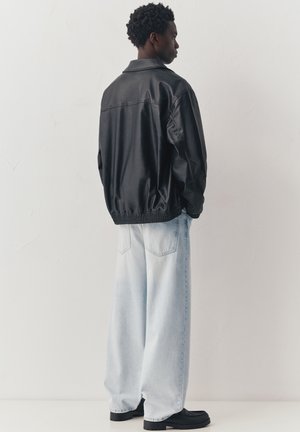 Mladi moški stoji bok, oblečen v črno usnjeno jakno, svetlo modre široke kavbojke in črne čevlje ob beli steni.