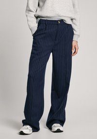 Pantalones a rayas azul marino hechos de una tela suave, con una cintura alta, bolsillos frontales y un cierre de botón. Combinados con una sudadera gris y zapatillas.