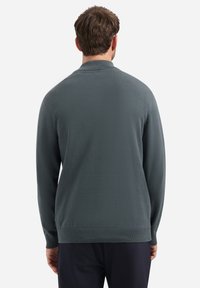 Man in een donkergrijze lange mouwen trui en zwarte broek, staand met de rug naar de camera tegen een effen witte achtergrond.