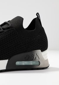 Talons de baskets en tricot noir avec semelle à coussin d'air intégrée et languette de traction sur une surface blanche sur fond neutre.