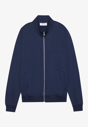 Tumes sinine jakksüda puuvilla segust, millel on kõrge kaelus, küljetaskud, soonikkud ja alläär, delikaatse tekstuuri ning minimalistliku disainiga.