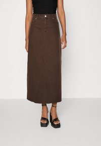 Falda maxi de denim marrón con costuras frontales, bolsillos laterales y corte recto. Llevada con sandalias de plataforma negras con tiras.