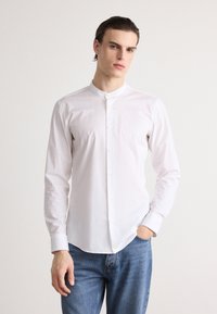 White button-up shirt with a band collar, long sleeves, and a smooth texture, paired with blue denim jeans.