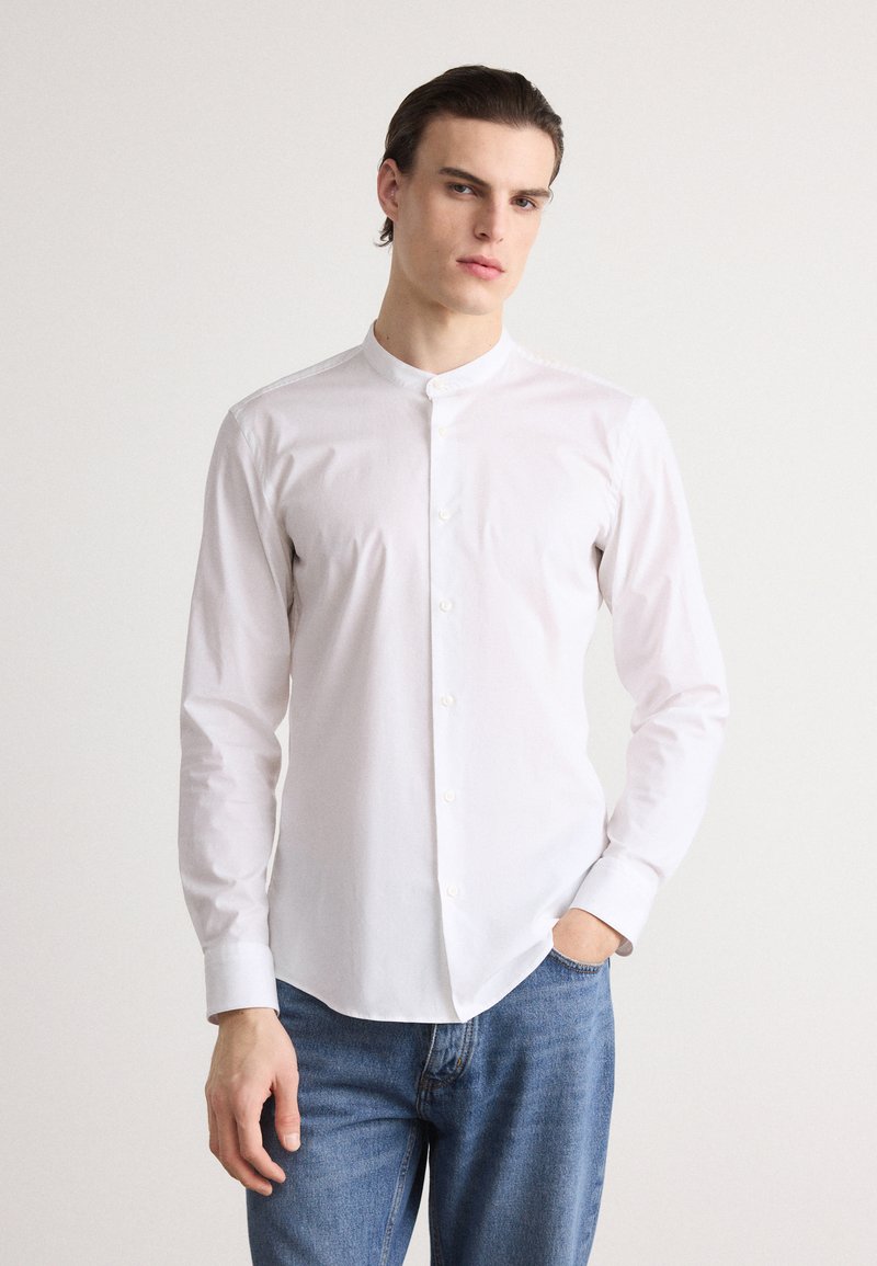 White button-up shirt with a band collar, long sleeves, and a smooth texture, paired with blue denim jeans.