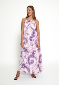 Robe maxi tie-dye violette avec des fines bretelles, un décolleté volanté et un tissu fluide présentant des motifs circulaires. Fond clair uni.