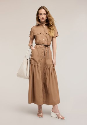 Jonge vrouw draagt een beige midi-jurk met knopen en geplooide rok, zwarte riem, witte sleehakken, draagt een grote witte boodschappentas.