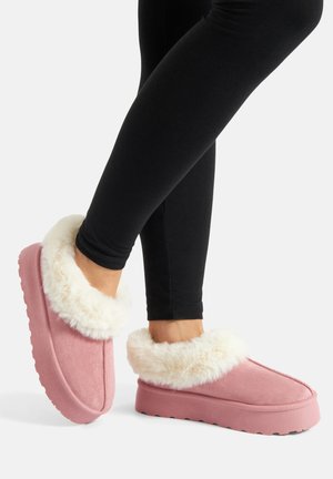 Feet wearing pink slip-on slippers with thick white fur lining and thick soles, paired with black leggings on a white background.