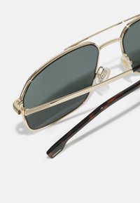 Goldene Metallsonnenbrille mit grauen Gläsern, versehen mit einem Doppelsteg-Design und schmal zulaufenden Bügeln im Schildpatt-Muster, die zu einem rechteckigen Ende führen.