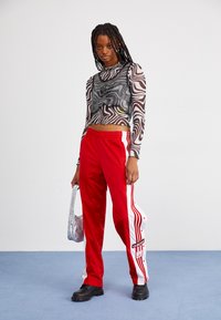 Calças de treino vermelhas com riscas brancas e a marca Adidas, combinadas com uma blusa transparente em preto e branco com padrão de zebra, e sapatos pretos slip-on.