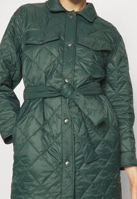 Chaqueta verde acolchada con patrón de diamantes, con una parte delantera abotonada, dos bolsillos en el pecho y una cintura ajustada con cinturón. Tejido suave y brillante.